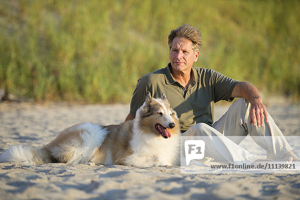 Kaukasischer Mann sitzt mit Hund am Strand