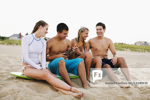 Teenager sitzen auf einem Surfbrett und spielen Ukulele am Strand