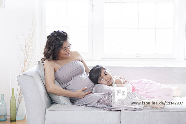 Hispanic girl listening to belly of pregnant mother on sofa