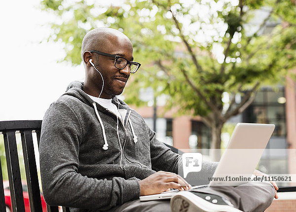 Schwarzer Mann mit Laptop und Kopfhörern auf einer Bank in der Stadt