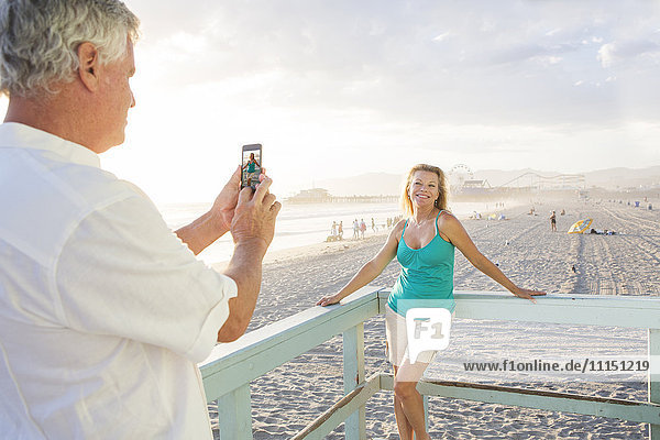 Caucasian man taking photograph of wife on boardwalk at beach
