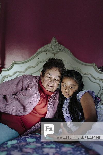 Hispanic grandmother and granddaughter using digital tablet on bed