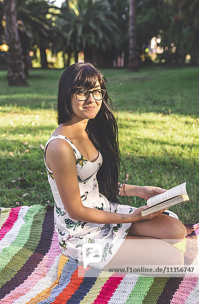 Young woman sitting on blanket in park holding a book