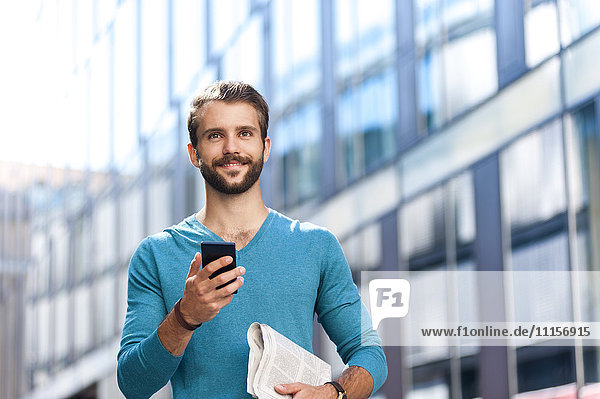 Lächelnder junger Mann mit Handy und Zeitung in der Stadt