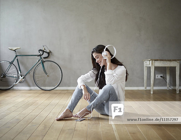 Smiling woman sitting on wooden floor at home listening music with headphones