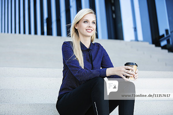 Lächelnde blonde Geschäftsfrau sitzt auf der Treppe mit Kaffee zum Mitnehmen