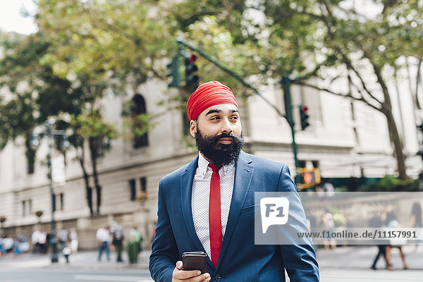 Indischer Geschäftsmann  der die Straße in Manhattan überquert und ein Smartphone hält.