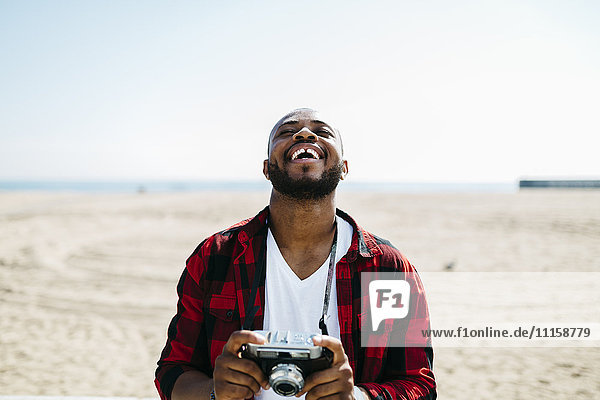 Lachender Mann hält eine altmodische Kamera in Strandnähe.