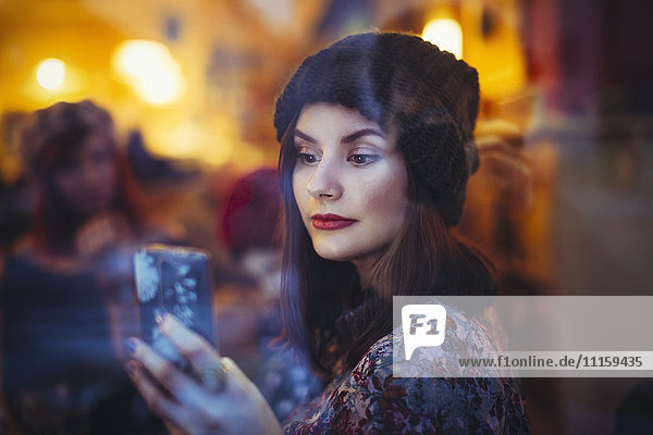 Porträt einer jungen Frau mit Smartphone hinter der Fensterscheibe einer Kneipe am Abend