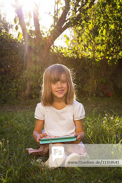 Portrait of smiling little girl sitting on a meadow with tablet