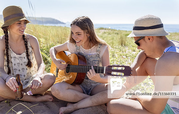 Lächelndes Teenagermädchen beim Gitarrespielen für ihre Freunde am Strand