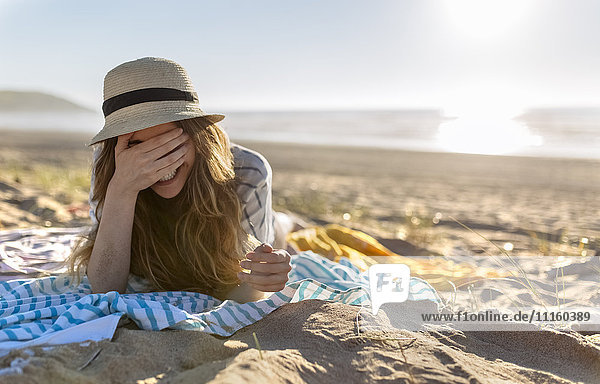 Portrait of smiling teenage girl lying on the beach covering face with her hand