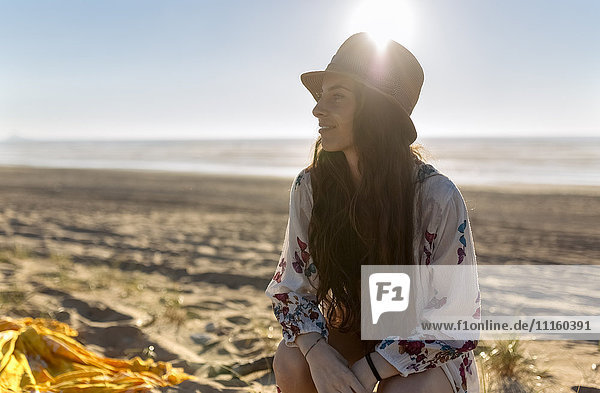 Lächelnde junge Frau sitzt am Strand bei Gegenlicht