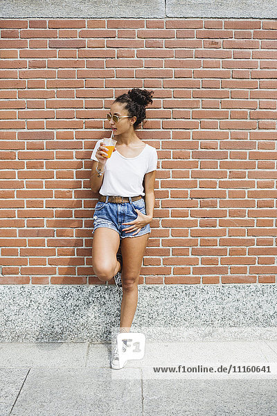Young woman leaning against a brick wall drinking orange juice