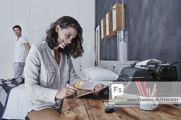 Smiling woman shopping online in bedroom with husband in background