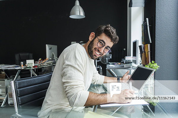 Porträt eines lächelnden jungen Mannes  der mit einem Laptop am Schreibtisch in einem modernen Büro arbeitet.