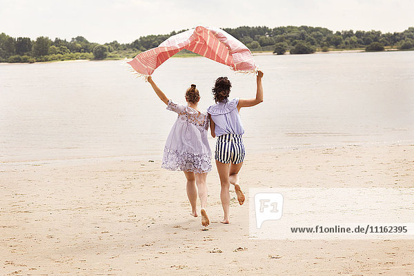 Rückansicht von zwei Freunden  die Seite an Seite auf dem Strand laufen und das Tuch über ihren Köpfen halten.
