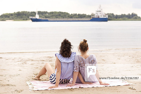 Back view of two friends sitting side by side on the beach watching a ship