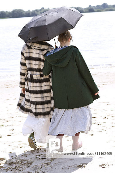Rückansicht von zwei Freunden  die Seite an Seite mit einem Regenschirm am Strand spazieren gehen.