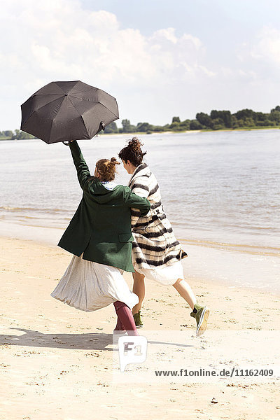 Rückansicht von zwei Freunden  die Seite an Seite am Strand mit einem Regenschirm laufen.