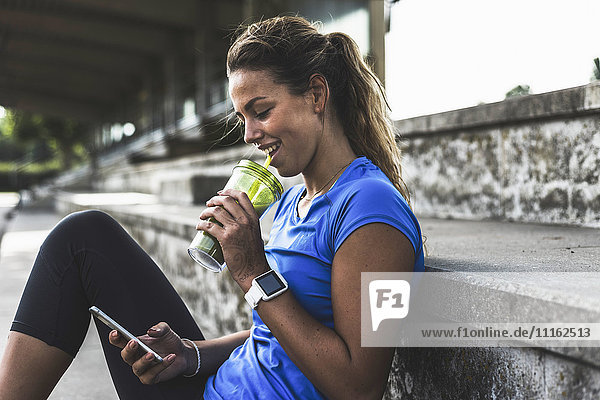 Sportive young woman sitting on grandstand with cell phone and drinking mug