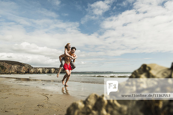 Fröhlicher junger Mann trägt Freundin Huckepack am Strand