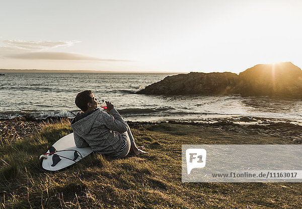 Frankreich  Bretagne  Halbinsel Crozon  Frau bei Sonnenuntergang an der Küste sitzend mit Surfbrett