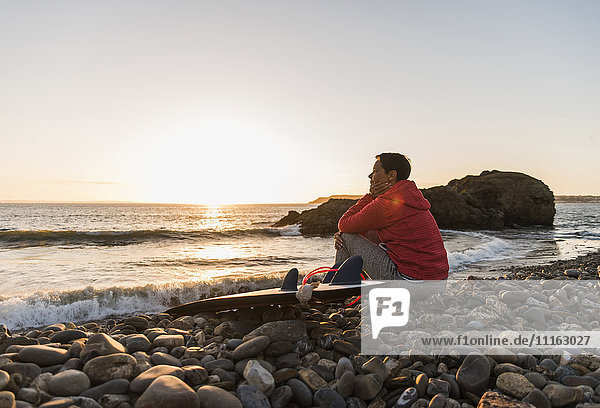 Frankreich  Bretagne  Halbinsel Crozon  Frau sitzt am steinigen Strand bei Sonnenuntergang mit Surfbrett