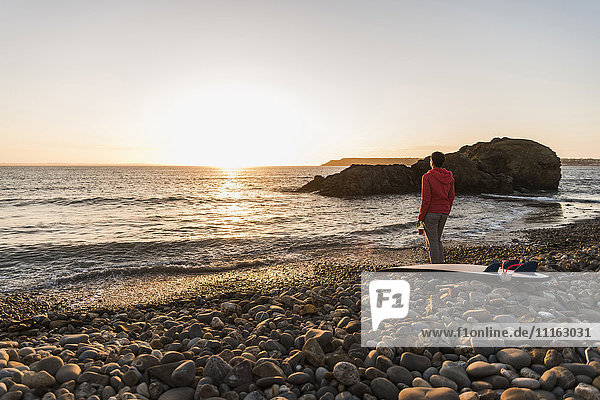 Frankreich  Bretagne  Halbinsel Crozon  Frau steht am steinigen Strand bei Sonnenuntergang mit Surfbrett