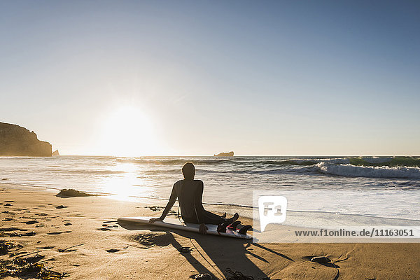 Frankreich  Bretagne  Halbinsel Crozon  Frau sitzt am Strand bei Sonnenuntergang mit Surfbrett