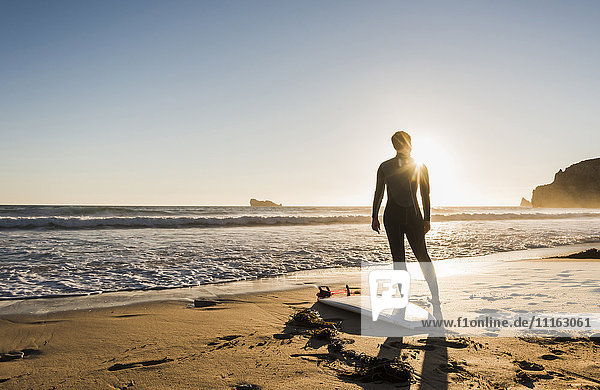 Frankreich  Bretagne  Halbinsel Crozon  Frau am Strand stehend bei Sonnenuntergang mit Surfbrett