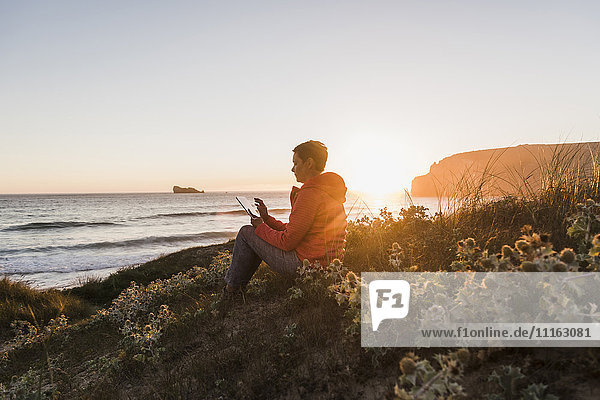 Frankreich  Bretagne  Halbinsel Crozon  Frau bei Sonnenuntergang an der Küste sitzend mit Tablette