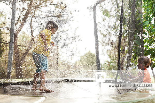Junge springt auf Trampolin  während sein kleiner Bruder ihn mit Wasser bespritzt.