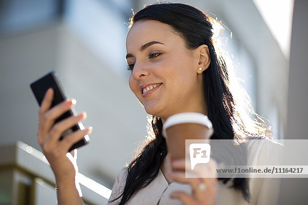 Lächelnde Frau mit Kaffee zum Mitnehmen beim Blick aufs Handy