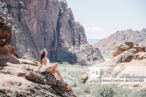 Junge Frau im weißen Badeanzug  die auf einem Felsen auf dem Canyonboden sitzt  mit Klippen und Gipfeln in der Ferne.
