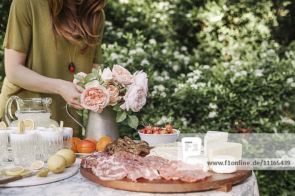 Frau  die an einem Tisch in einem Garten steht  eine Vase mit rosa Rosen  Getränken  einer Schale mit Kirschen und einem Holzbrett mit Aufschnitt und Käse.