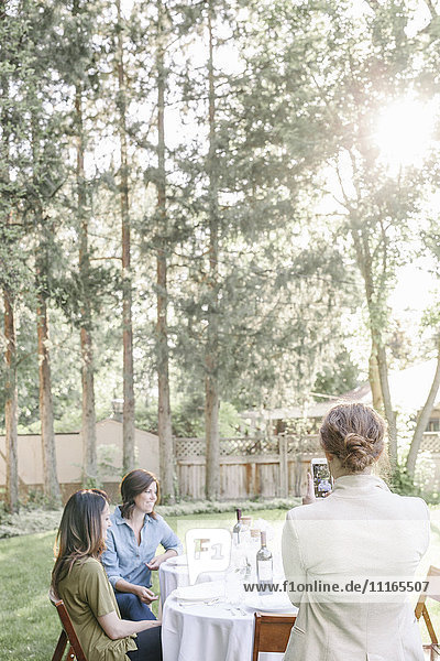 Eine Frau fotografiert mit ihrem Mobiltelefon eine Gruppe von Frauen  die an einem Tisch in einem Garten sitzen.