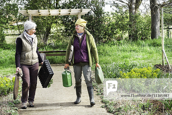 Zwei Frauen gehen einen Weg entlang  die Knieschützer  Gießkanne und Pflanztabletts tragen.