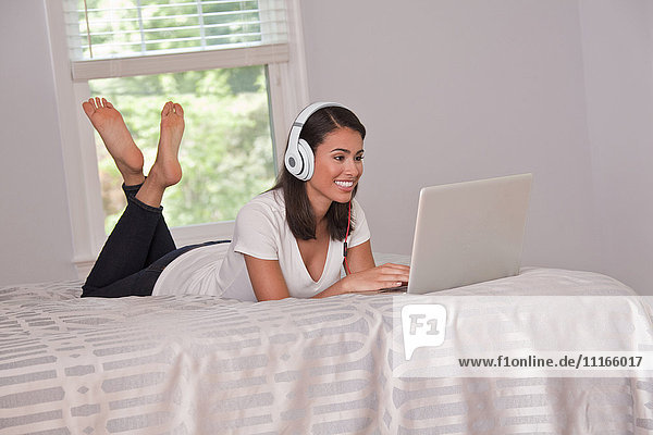 Hispanische Frau liegt auf dem Bett und hört mit Kopfhörern auf ihren Laptop