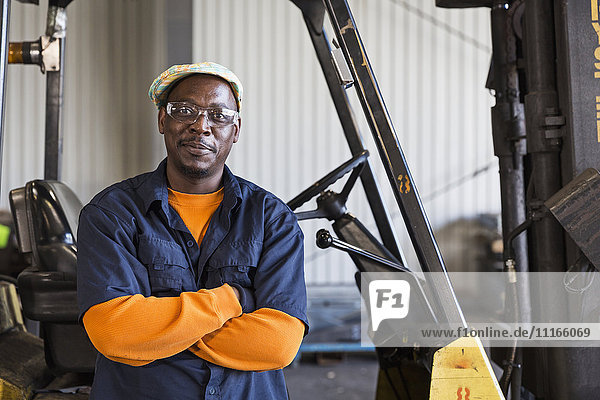 Lächelnder schwarzer Arbeiter posiert in der Nähe eines Gabelstaplers in einer Fabrik