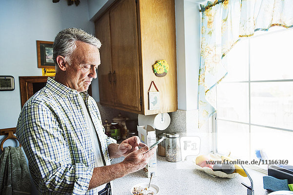 Älterer Mann steht in einer Küche und benutzt ein Mobiltelefon.