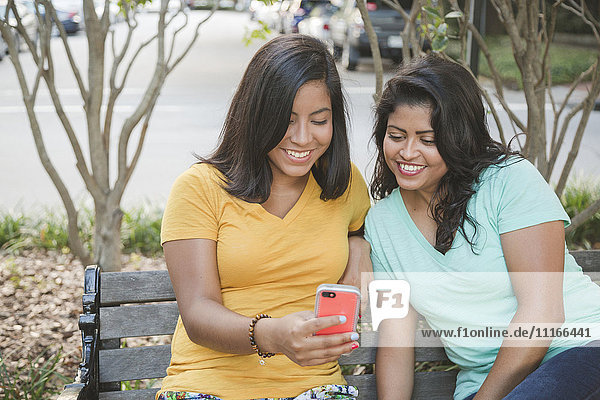 Hispanische Mutter und Tochter benutzen ein Handy auf einer Parkbank