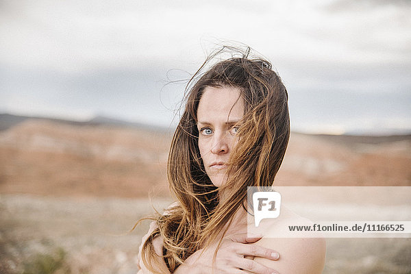 Eine Frau mit langen braunen Haaren steht in einer Wüstenlandschaft. Verschränkte Hände und nackte Schultern.
