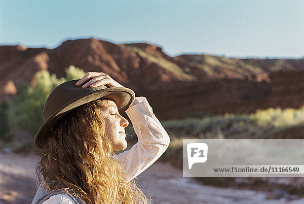 Eine Frau mit Hut steht im offenen Raum mit Bergen  der Sonne zugewandt.