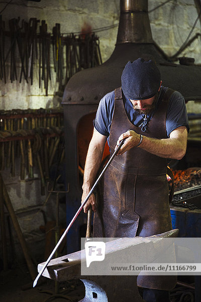 Ein Schmied in einer Lederschürze hält ein dünnes Stück Metall gegen einen Amboss in einer Werkstatt.