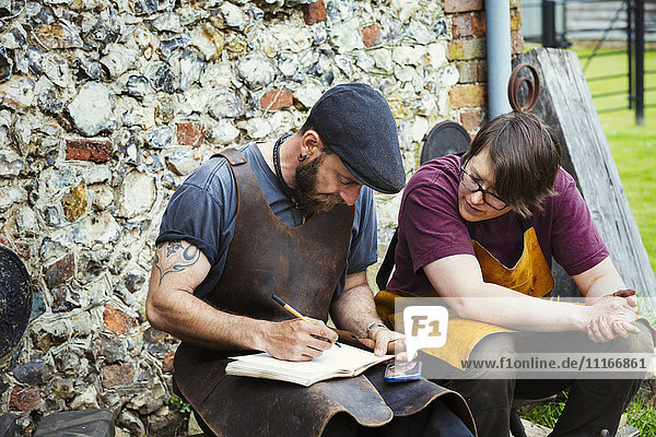 Two men wearing aprons writing into a notebook sat in a garden.