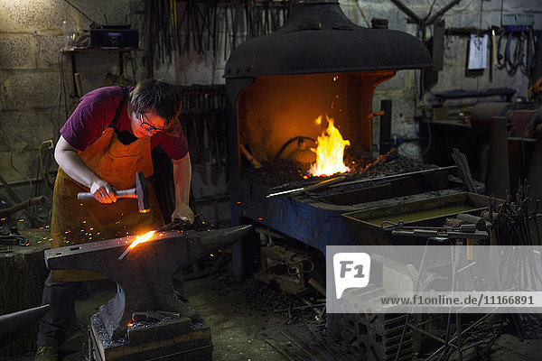 Ein Schmied schlägt rotes  glühendes Metall auf einen Amboss in einer Werkstatt.