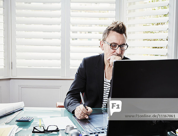 Ein Mann mit Brille sitzt an einem Schreibtisch in einem Büro und schaut auf einen Computerbildschirm.