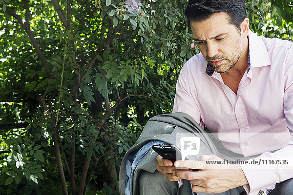 Businessman wearing a grey suit and pink shirt sitting outdoors  using his cell phone.