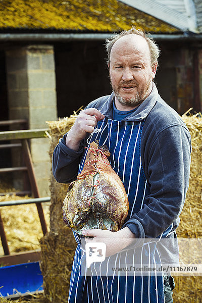 Butcher wearing a striped blue apron  standing outdoors  holding a large cured ham.